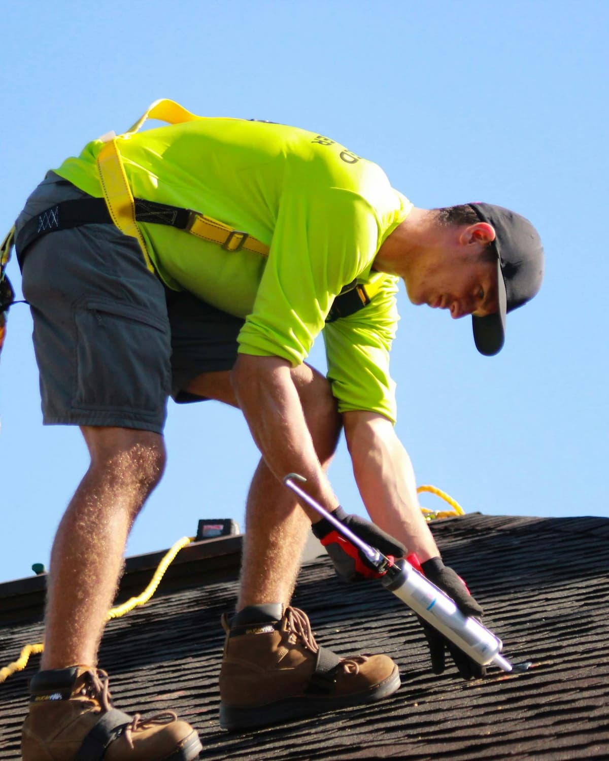 Roofer in a high-visibility shirt and safety harness applying caulk to shingles on a roof under a clear blue sky
